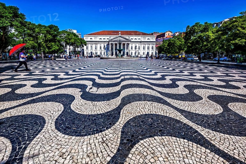 Wavy black and white patterned pavement in a sunny urban square with people and classic building in background, Lisbon