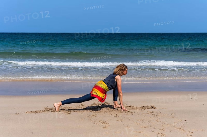 A woman doing yoga on a sandy beach by the sea. Ashwa Sanchalanasana or Sprinter, variant. Treen Cove, Cornwall, England, Great Britain