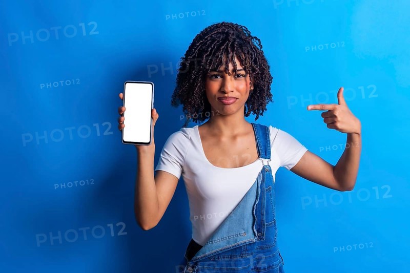 Young woman showing a smartphone with blank white screen and pointing at it with index finger, promoting mobile app or website