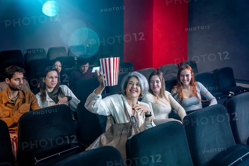 Mature woman enjoying a film in a movie theater, holding a popcorn bucket, surrounded by other people