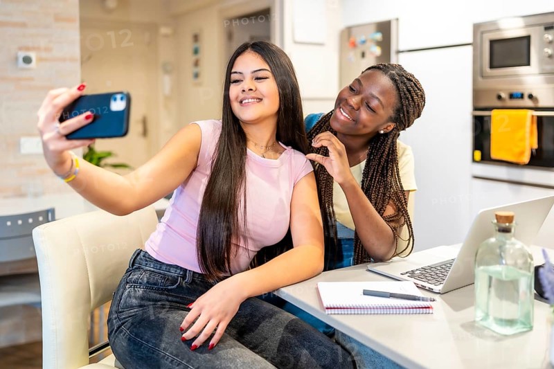 Two happy teenage girls taking a selfie with a smartphone while doing homework in a modern kitchen