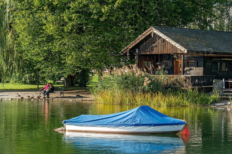 Wooden hut with common reed (Phragmites australis), covered boat, two woman on a park bench, shore of Lake Schliersee, spa garden Markt Schliersee, Upper Bavaria, Bavaria, Germany, Europe