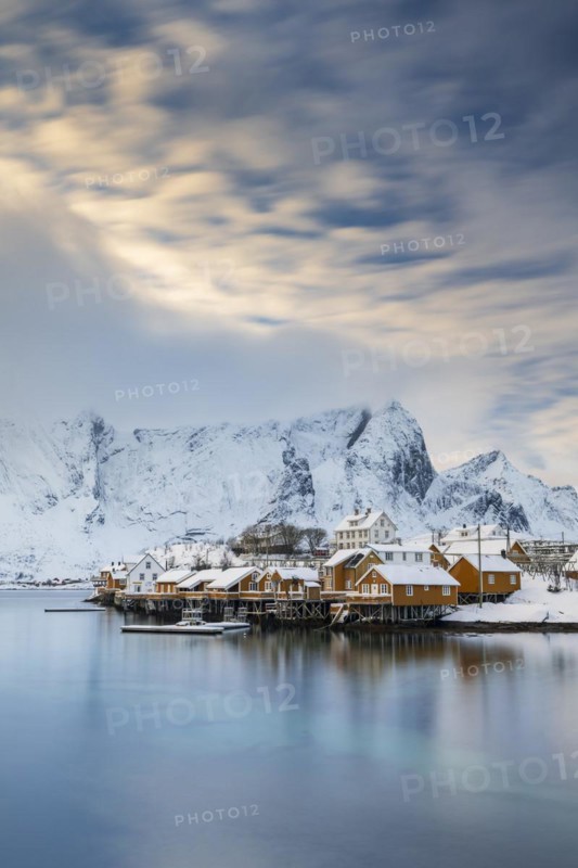 Traditional yellow rorbuer huts on the island of Sakrisøy, snow-capped mountains in the background, Sakrisoy, Reine, Lofoten, Norway, Europe