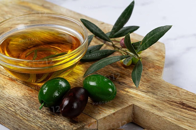 A glass bowl filled with rich olive oil sits on a wooden cutting board alongside fresh green and black olives, complemented by olive leaves, creating a rustic culinary setting