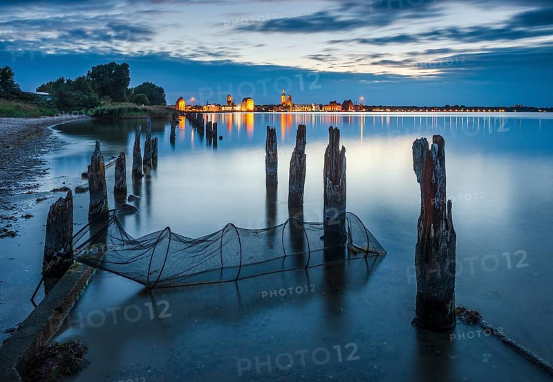 View at dusk over the Strelasund to the illuminated town of Stralsund with its churches and the warehouses in the harbour, in front old groynes and a fishing net, Stralsund, Mecklenburg-Vorpommern, Germany, Europe