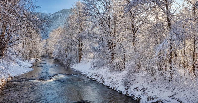 A calm river flows through a snowy winter landscape, surrounded by snow-covered trees and mountains in the background, Leitzach