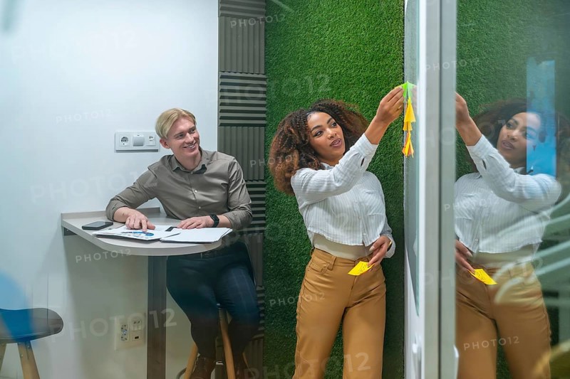 Businesswoman attaching sticky notes on a glass wall while brainstorming with a colleague in a modern coworking space