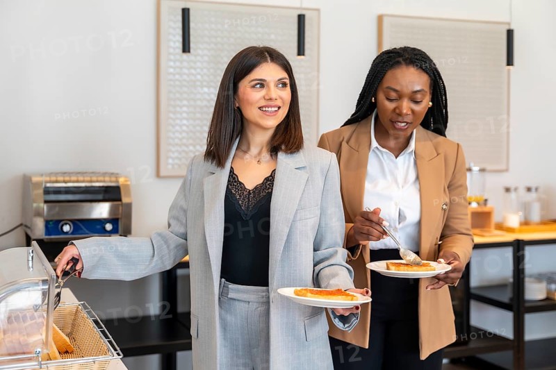 Two businesswomen are enjoying breakfast at a hotel buffet, adding jam to their toast during a business trip
