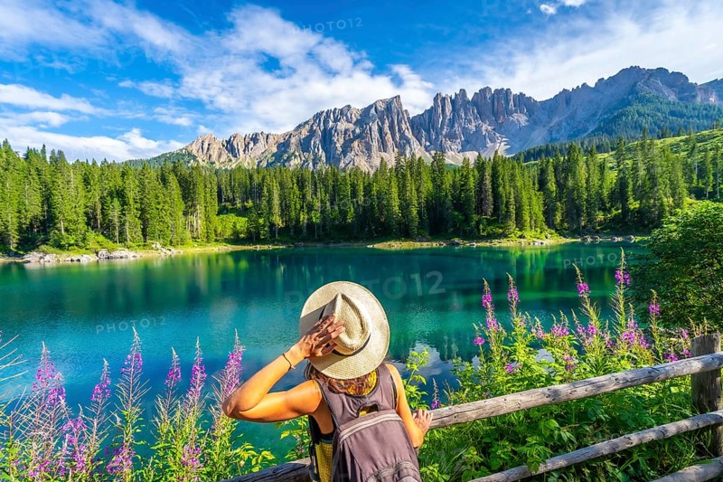 Woman with backpack and hat admiring the turquoise waters of lake carezza with the latemar mountain range in the background, dolomites, italy