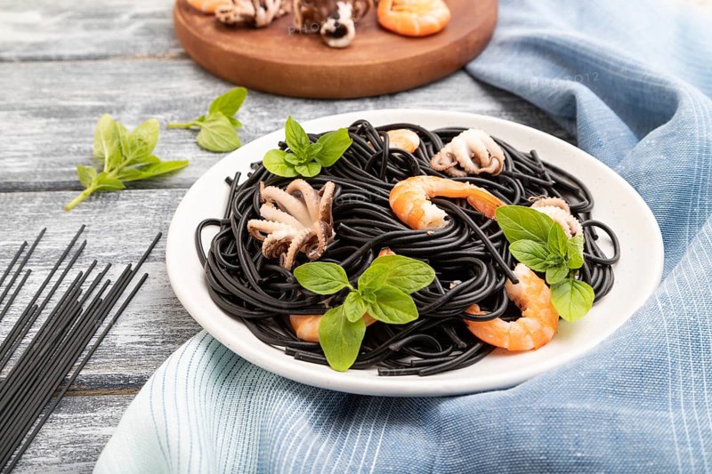 Black cuttlefish ink pasta with shrimps or prawns and small octopuses on gray wooden background and blue textile. Side view, close up