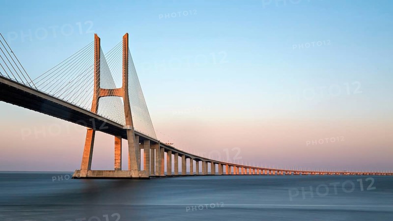 A long cable-stayed bridge spans over water under a gradient sunset sky, Lissabon, Vasco Da Gama-Brücke