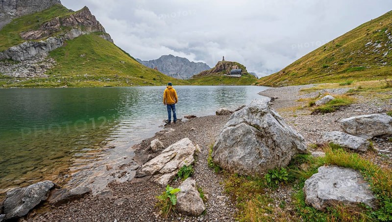 Mountaineer in yellow jacket at the mountain lake, view over the Wolayersee lake to the alpine club hut Wolayerseehütte, cloudy mountain landscape, Carnic Alps, Carnic High Trail, Carinthia, Austria, Europe