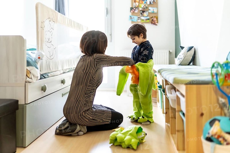 Mother assisting her son in getting dressed in a dinosaur costume, fostering imaginative play in a cozy bedroom setting