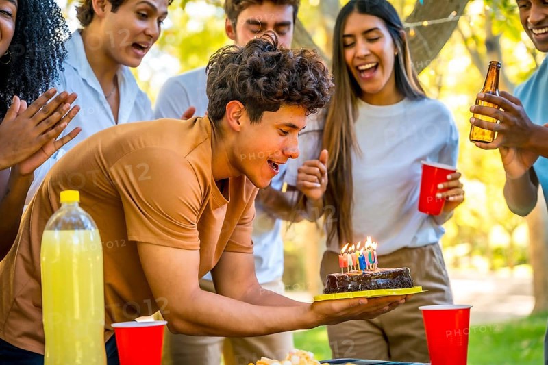 Happy young man with with birthday cake celebrating with friends at park