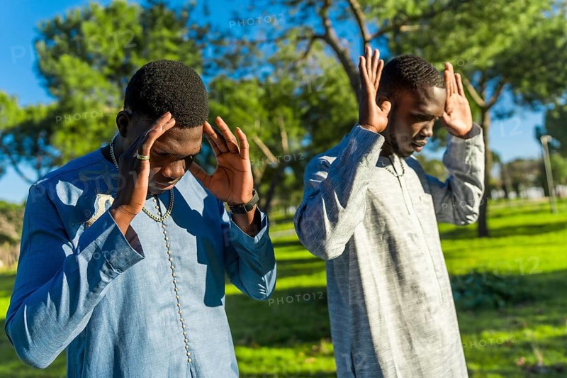 Two young senegalese men wearing traditional dashiki clothing are praying outdoors in a park, observing ramadan
