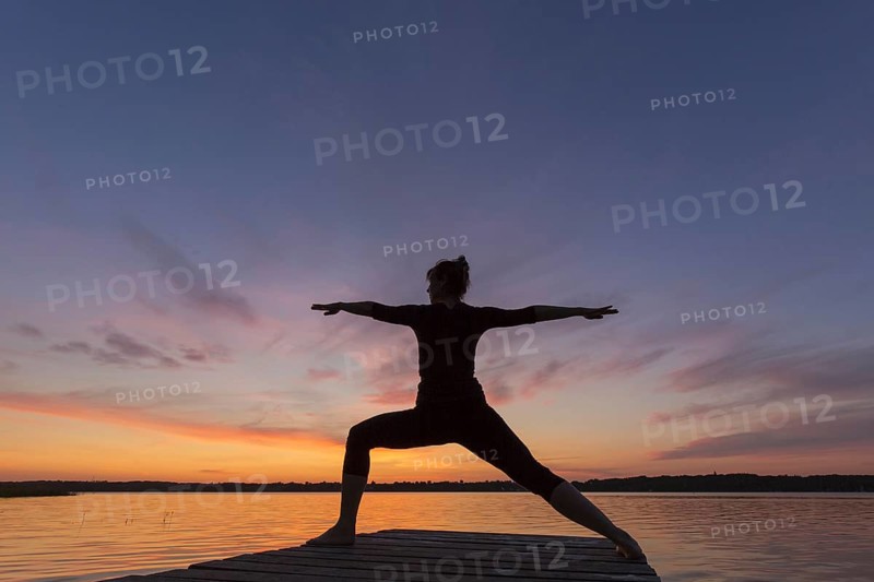 Woman practicing yoga posture Virabhadrasana II, Warrior Pose, lunging standing asana on jetty at lake at sunset