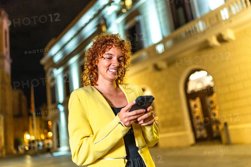 Smiling cheerful woman in yellow clothes and curly hair using smartphone walking along city at night