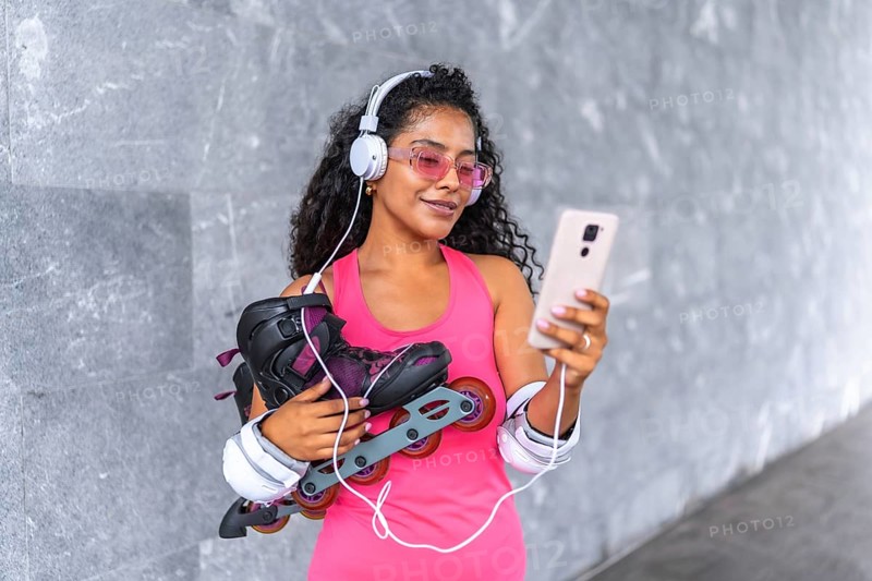 Latin beauty woman with inline skates listening to music with mobile phone and headphones at urban park