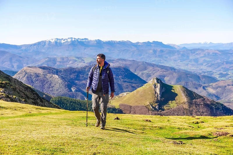 Male hiker walking on grassy mountain slope with trekking poles, enjoying scenic view of mount txindoki and surrounding peaks in gipuzkoa, basque country, spain
