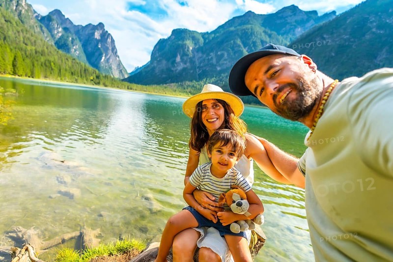 Happy family taking a selfie by the transparent water of lake dobbiaco with the dolomites in the background, enjoying summer vacation