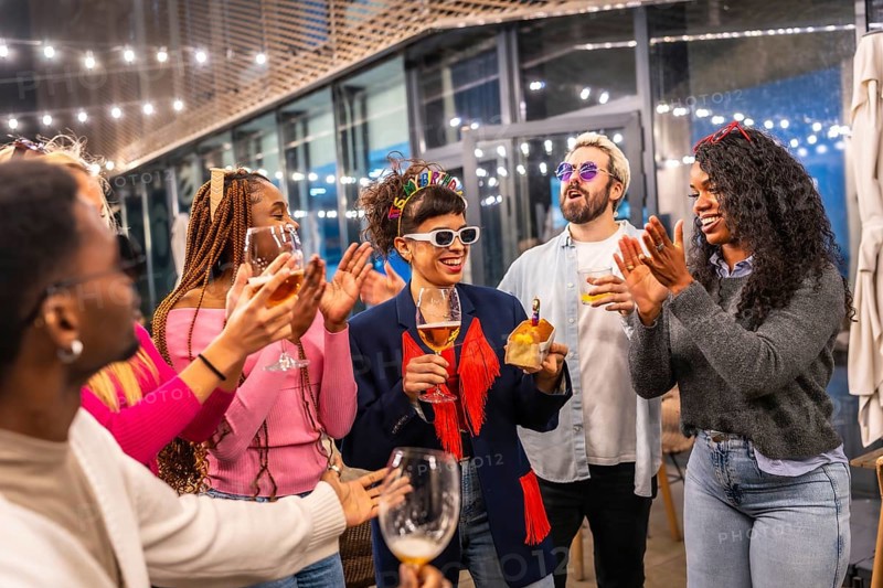 Multi-ethnic friends clasping and singing birthday song to a woman standing outside a night bar terrace