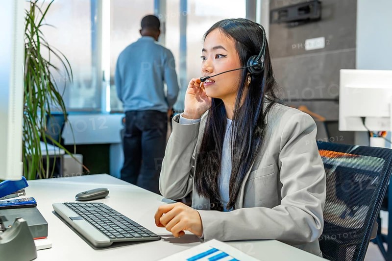 Young asian manager working diligently at a computer, wearing a headset in a sleek, modern coworking office environment