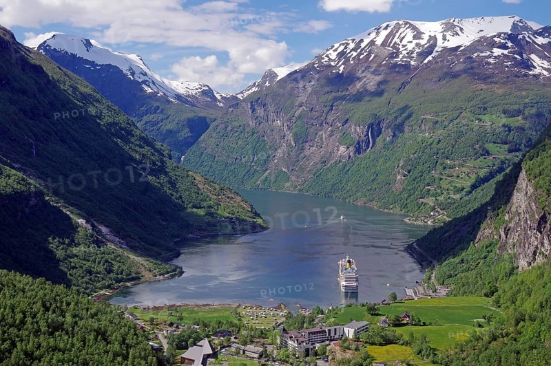 A fjord with a cruise ship surrounded by green mountains with snow-capped peaks under a blue sky with white clouds, Geirangerfjord, Fjordland, Norway, Europe