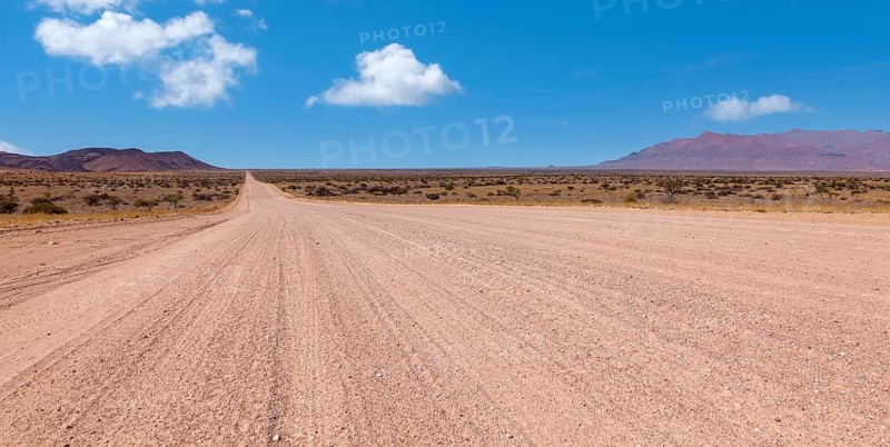 An endless desert road under a blue sky with a wide, dry landscape and mountains in the background, gravel road D2315, Namibia, Africa
