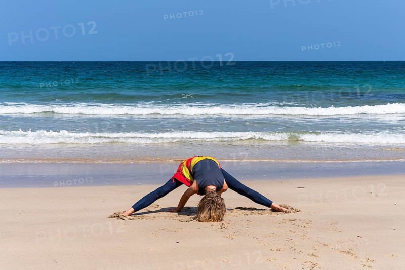 A woman doing yoga on a sandy beach by the sea. Prasarita Padottasana or forward bend with legs apart. Treen Cove, Cornwall, England, Great Britain