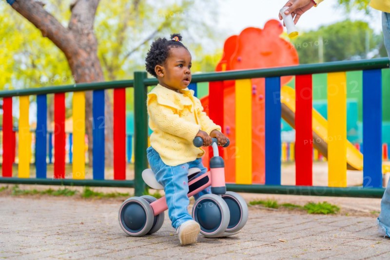 Full length photo of a cute african baby girl riding a toy bike in a park