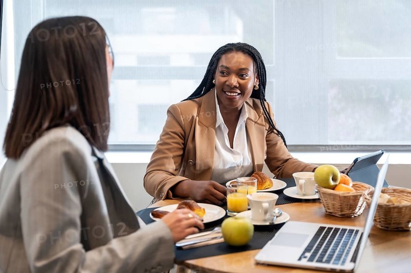 Two businesswomen enjoying breakfast and engaging in conversation during their business trip, utilizing laptop and tablet
