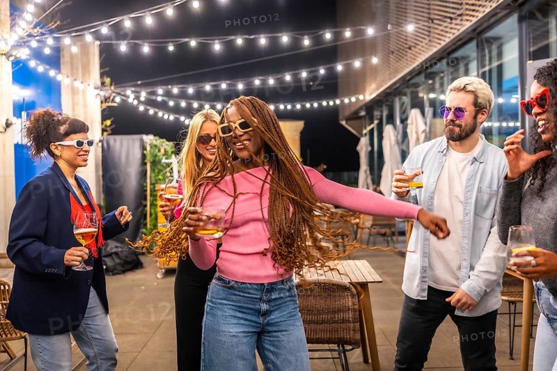 Happy modern african woman with long braided hair dancing with multi-ethnic friends at night party outdoors in the terrace of a bar