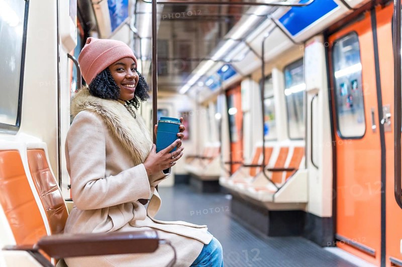 Smiling young black woman wearing winter clothes holding reusable coffee cup while riding subway train