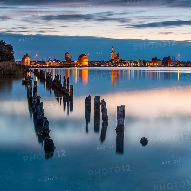 View at dusk over the Strelasund to the illuminated town of Stralsund with its churches and the warehouses in the harbour, old groynes in front, Stralsund, Mecklenburg-Western Pomerania, Germany, Europe