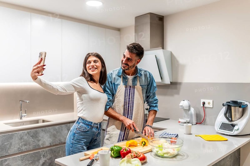 Young couple taking selfie with smartphone while preparing healthy meal together in modern kitchen
