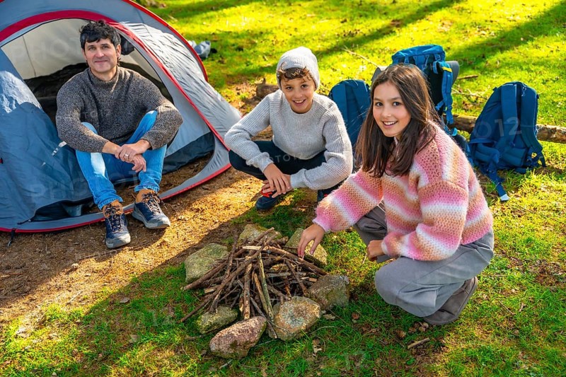 Caucasian father and two sons preparing a bonfire with dry branches in the forest next to camping tent