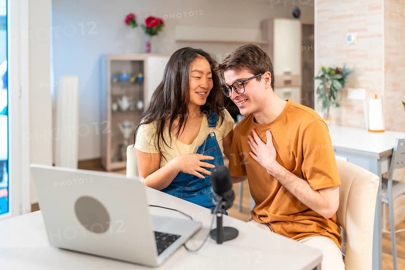 Young multiethnic couple sitting together at home, using a laptop and microphone to record their engaging audio podcast
