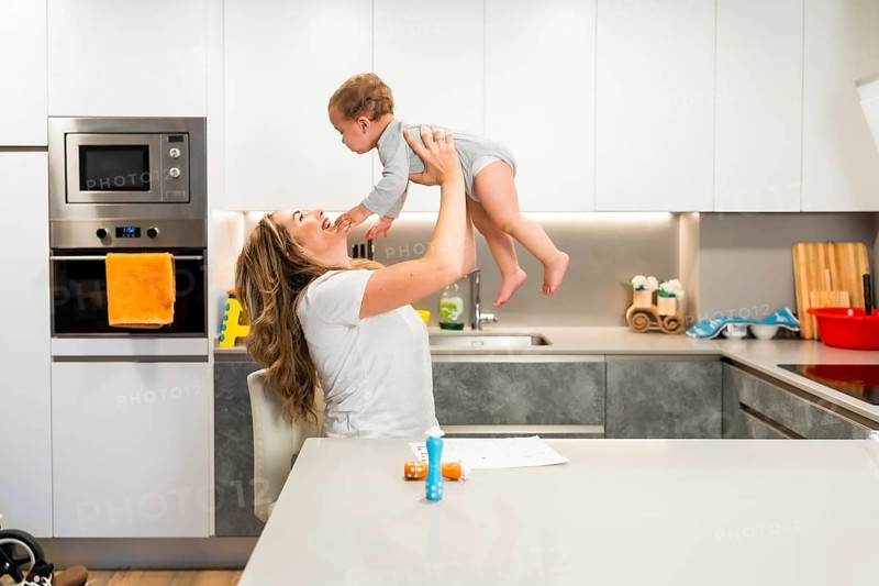 Happy mother playing with baby, lifting her in the air in a modern kitchen, enjoying time together
