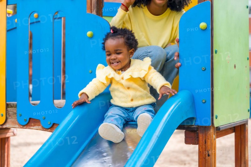 Cute african girl sliding in slide in a colorful playground next to her mother