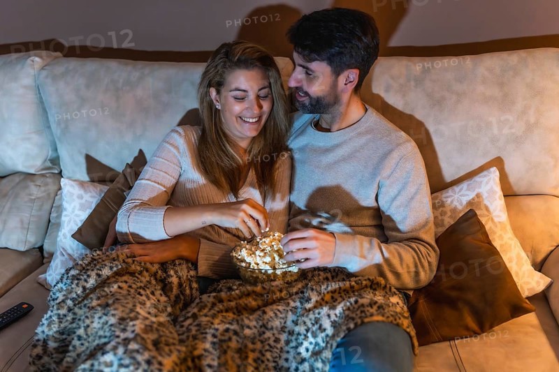 Romantic couple sharing a bowl of popcorn while watching a movie on the sofa at night, enjoying a cozy and intimate moment together