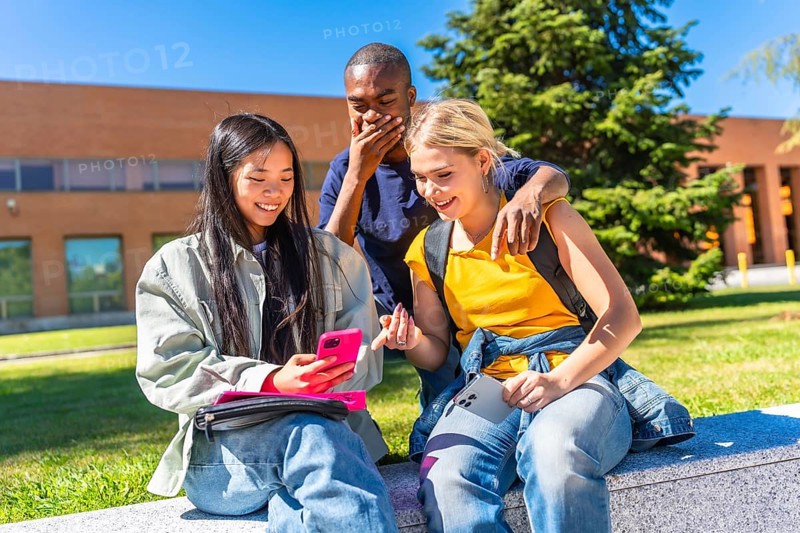Multiracial group of students using mobile and expressing surprise sitting in the campus