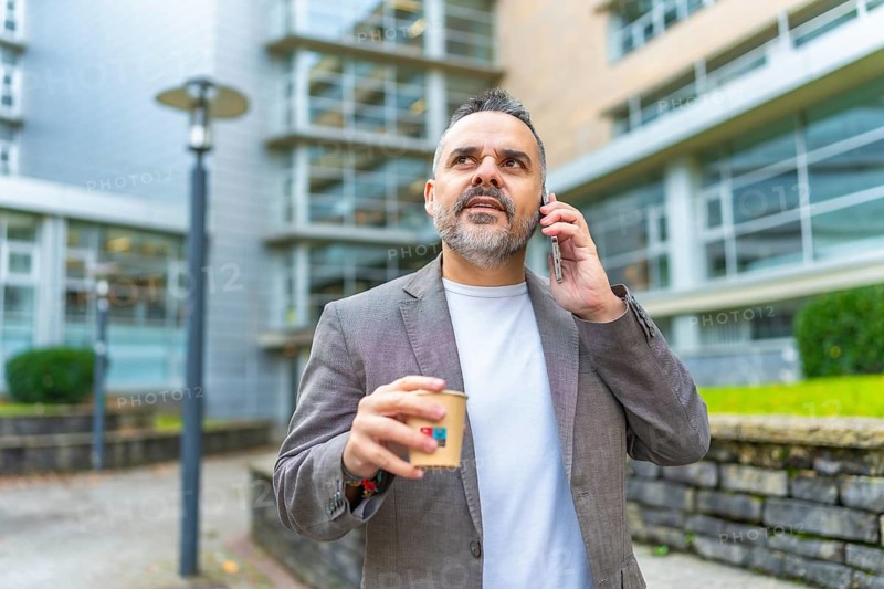 Mature caucasian businessman talking to the smartphone while drinking take away coffee outdoors