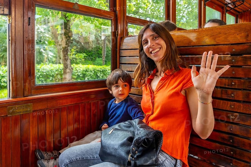 A woman and a child are sitting on a train. The woman is wearing an orange shirt and is waving at the camera