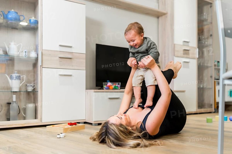Young mother lying on the floor lifting her baby in the air while playing together in the living room at home