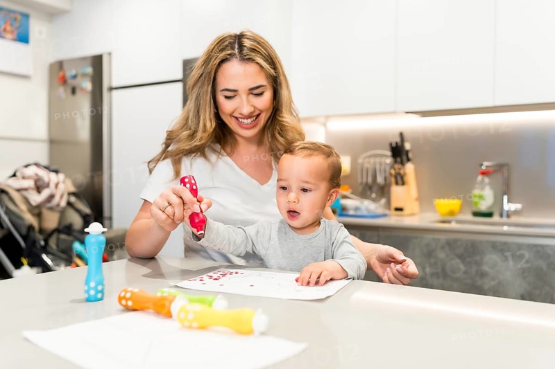 Smiling mother and baby boy having fun drawing together with colorful dot markers in a modern kitchen