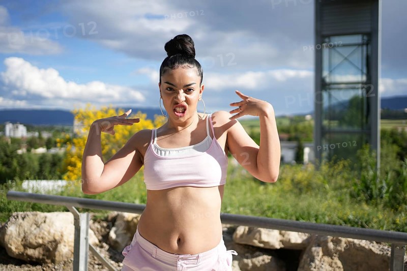 A woman is posing for a picture with her arms raised and a ponytail. She is wearing a pink top and shorts. Scene is energetic and confident