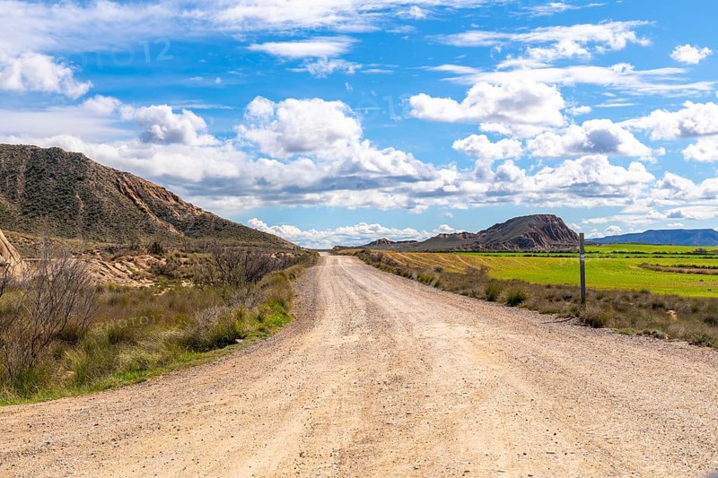 Scenic view of a gravel road stretching through the bardenas reales desert landscape under a bright blue sky with fluffy white clouds