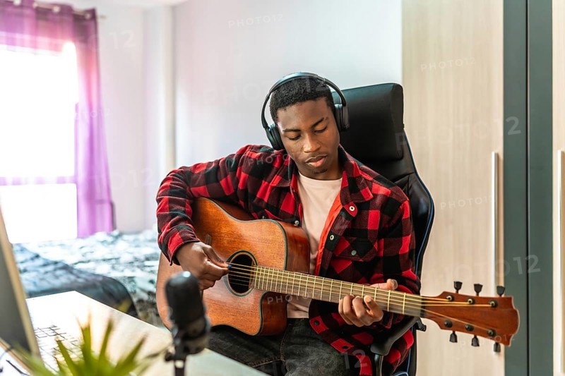 Young man playing acoustic guitar and composing music at home studio using headphones and microphone