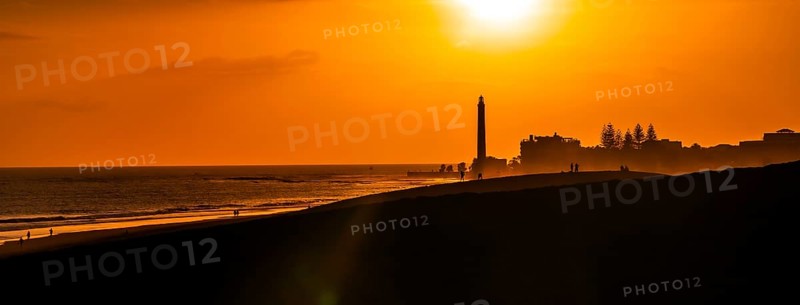 Tourists enjoying a beautiful sunset over maspalomas dunes in gran canaria, spain, with the iconic lighthouse silhouetted against the orange sky