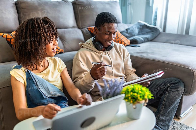 Two college students collaborating on a project, using a laptop and taking notes, demonstrating teamwork and focused learning in a comfortable home environment